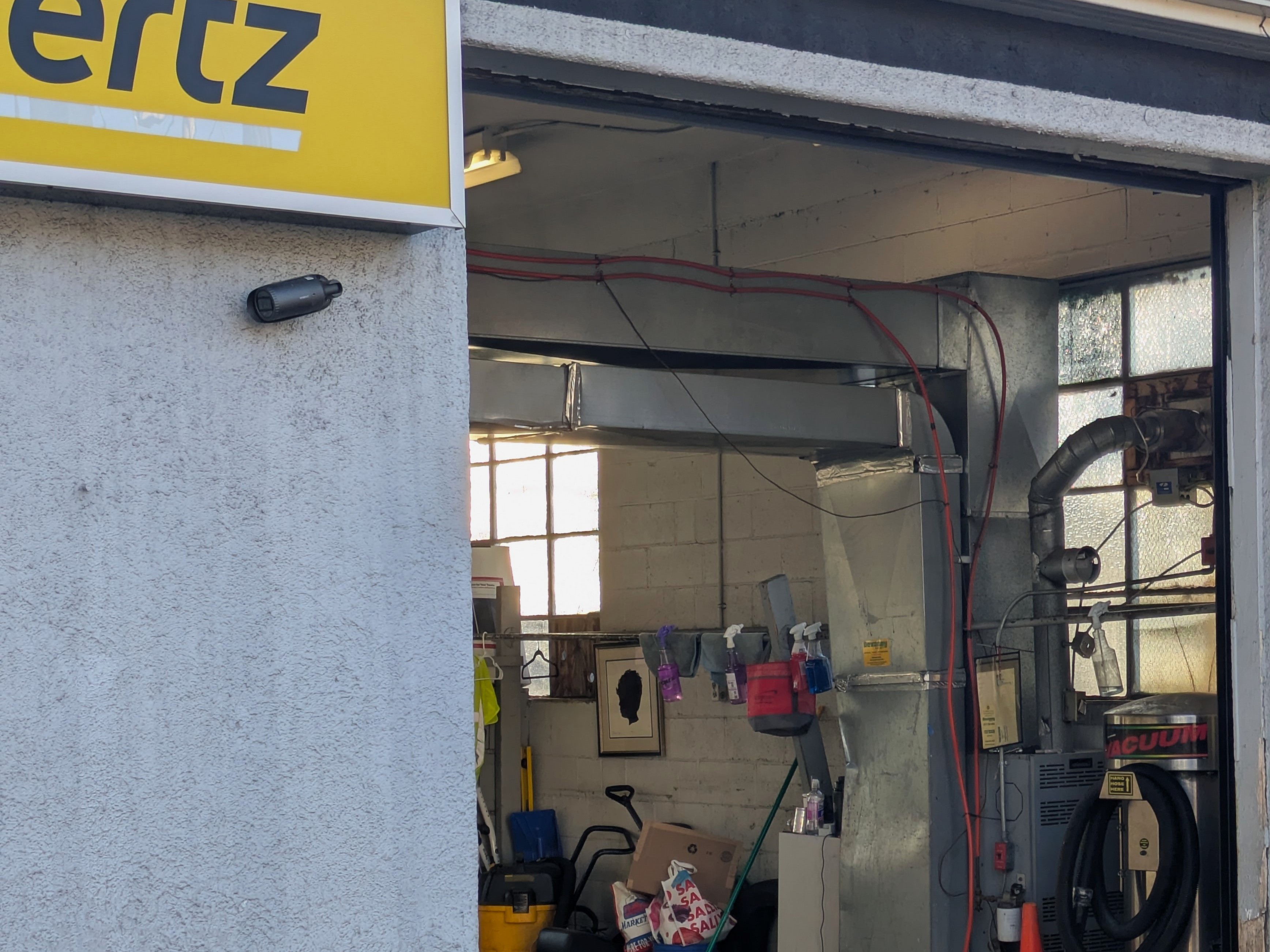 An open garage door. The inside is cluttered with car maintenance supplies. A framed paper silhouette of a child hangs on the cinderblock wall.