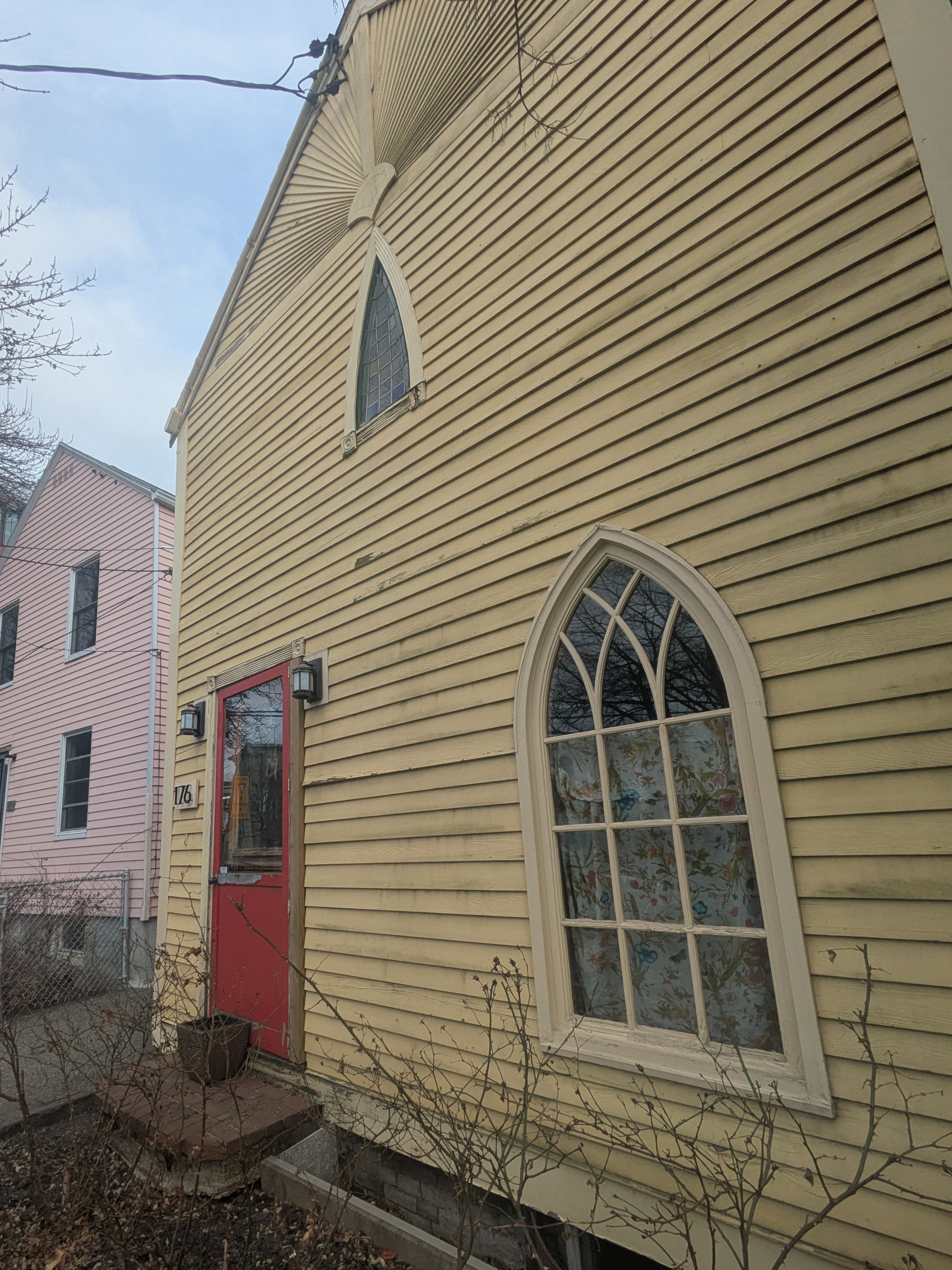 A butter yellow house with a red door. Next to the door is an arched, cathedral-like window with contemporary-seeming muntins. On the second story is another arched window, with historical-seeming leaded glass panes. The horizontal siding of the house turns into a fan-like pattern of rays across the gable.