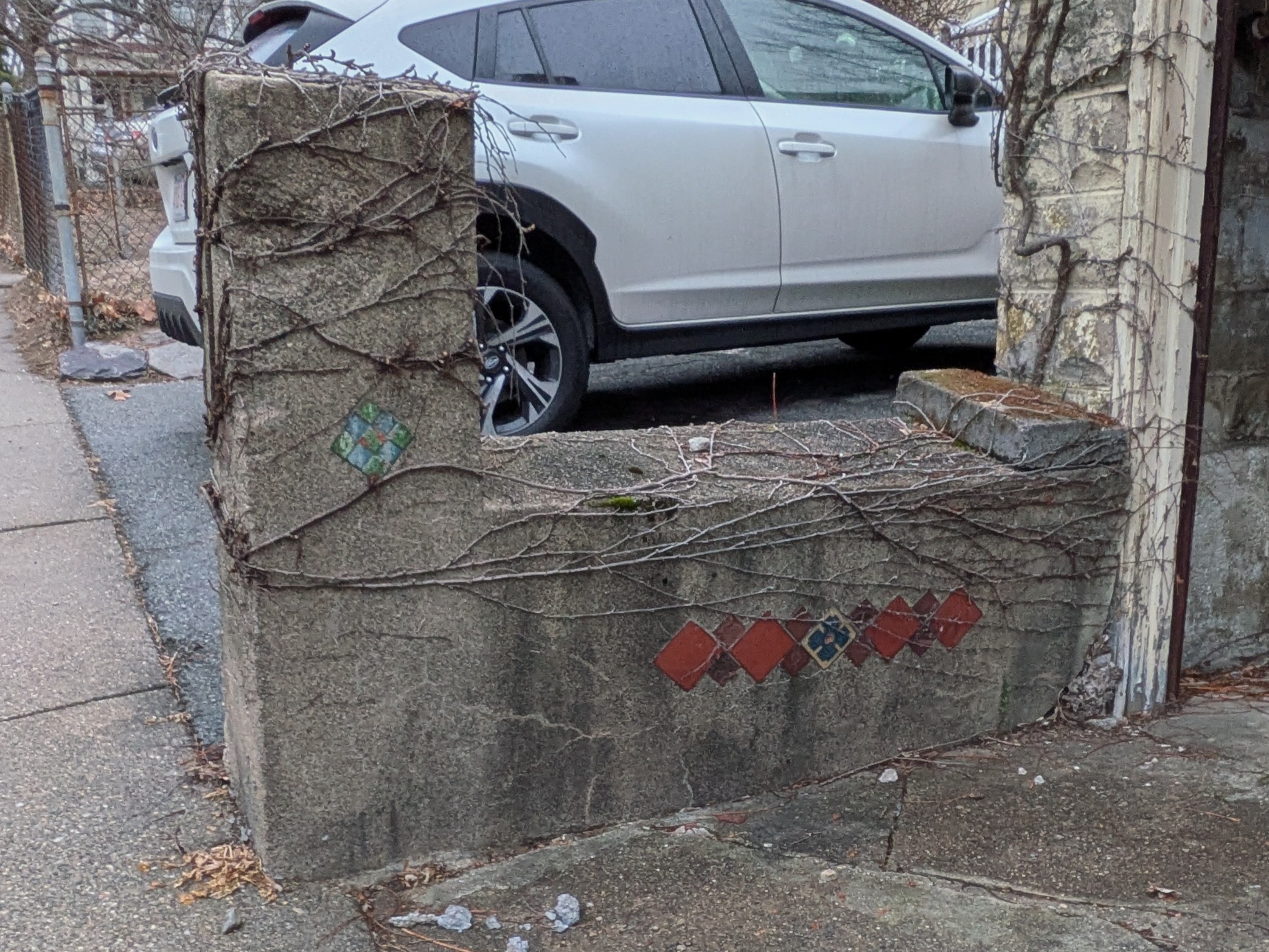 The cement sidewall of the driveway exiting the previous garage. A strip of red diamonds is tiled across it. The centermost tile looks like a blue four-petaled flower. 
