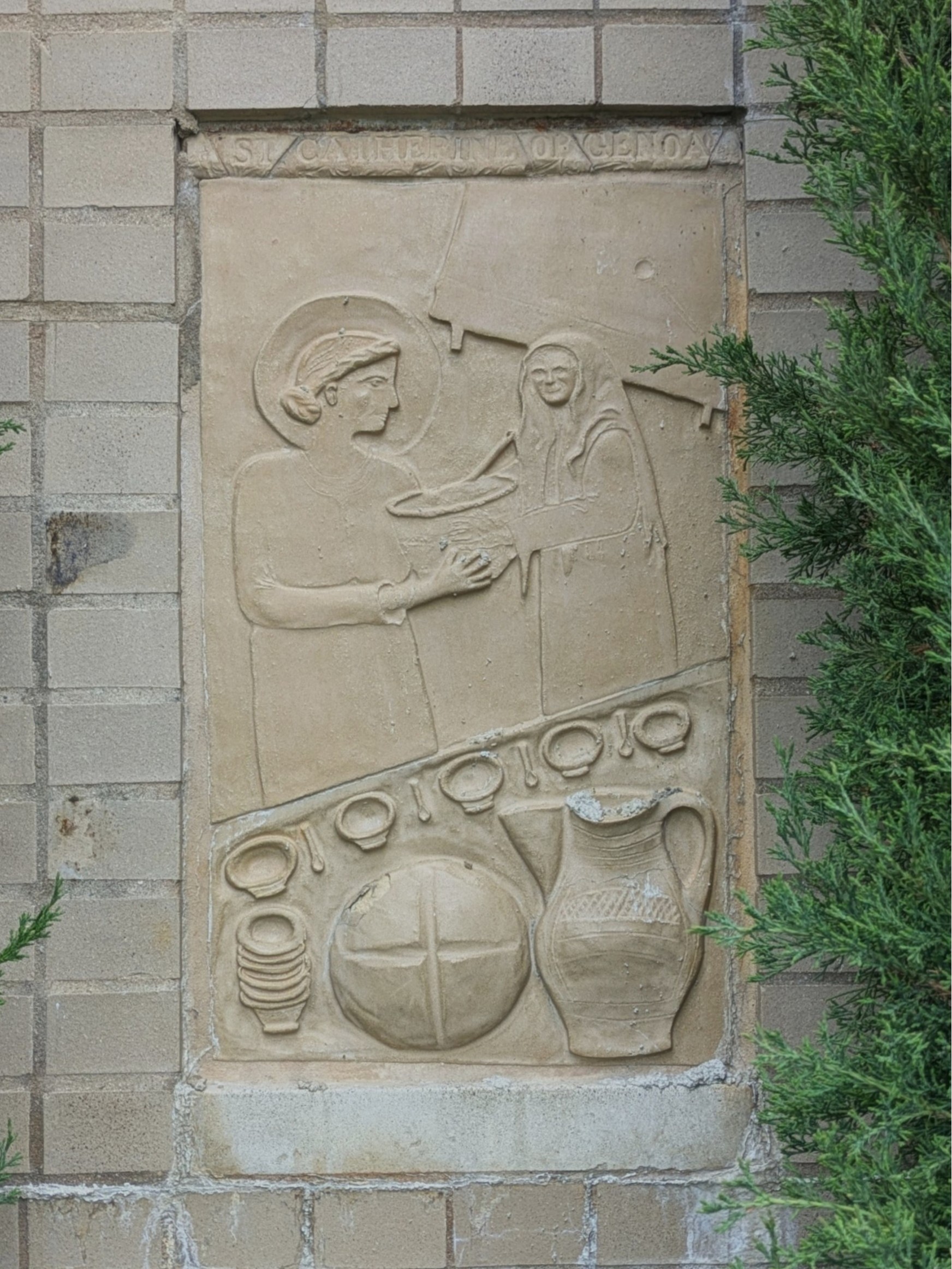 A clay carving of a woman standing in front of a table of food and dishware. She has a halo around her head, and is handing a bowl to a cloaked woman in the background. Behind them both is another table. The perspective of the whole image is oddly flat.